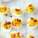 bacon cheddar deviled eggs on a white wooden board with bacon crumbles and paprika sprinkled around for decoration
