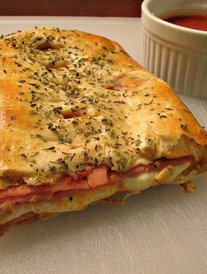 Photo of sliced Ham and Cheese Crescent Roll Stromboli on a white cutting board next to a white bowl of marinara sauce