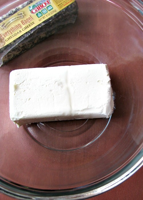 Picture of softened cream cheese in a clear bowl next to a bar of Cabot Cheese Everything Bagel cheddar cheese 