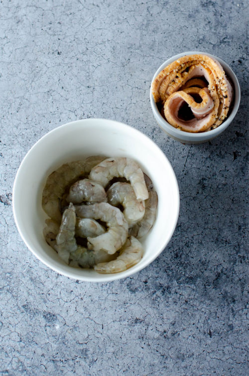 two bowls one with raw shrimp and one with raw bacon on a blue counter